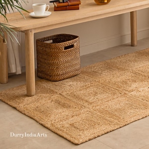 May include: A woven brown basket with a rectangular shape and cut-out handles sits on a natural fibre rug. The rug has a geometric pattern and is under a light-coloured wooden table. A cup and saucer are on the table.