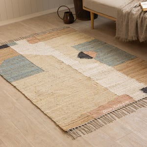 May include: A rectangular woven area rug featuring a geometric pattern in shades of tan, white, blue, and pink. The rug has a textured surface with fringe detailing on the short ends and the text DhurryIndiaArts printed in the bottom right corner.