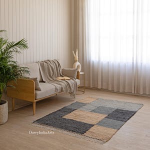 May include: A rectangular area rug with a geometric pattern in shades of gray, beige, and black. The rug has a fringed edge and is placed on a wooden floor. The rug is in a living room setting with a sofa and a window.