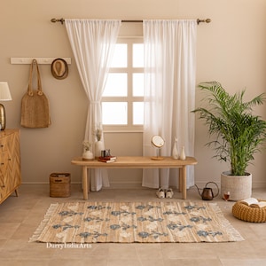 以下が含まれることがあります： 青と白の模様のジュートラグ、木製のベンチ、白いカーテンを備えたニュートラルな色調のインテリアシーン。織りのバッグと帽子が壁に掛かっています。鉢植えの植物と小さな鏡が装飾を加えています。