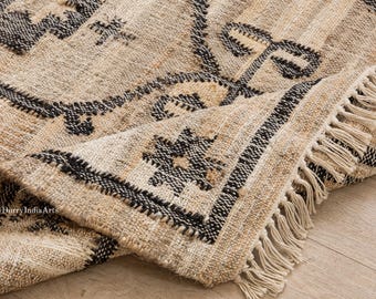 May include: Beige and black woven rug with a geometric pattern. The rug has a fringed edge and is made of a natural fiber material. The design features black embroidered details and a border.