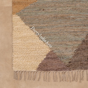 Op de afbeelding: Een geweven vloerkleed met een geometrisch patroon van crèmekleurige, beige, blauwgrijze en donkerbruine vezels. Het kleed heeft een getextureerd, geknoopt oppervlak en een franjerand aan de onderkant. De tekst DurryIndiaArts staat op het kleed.