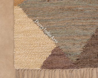 May include: A woven area rug featuring a geometric pattern with blocks of cream, tan, blue-gray, and dark brown fibers. The rug has a textured, knotted surface and a fringe edge along the bottom. The text DurryIndiaArts is printed on the rug.