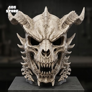 May include: A beige, bone-colored demon skull mask featuring prominent horns, sharp teeth, and intricate textured details. The text AEG STORE is visible in the upper left corner. The mask has a weathered, aged appearance with deep crevices and ridges.