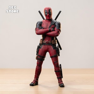 May include: A Deadpool action figure in a red and black suit, with two swords on its back and a utility belt. The figure has its arms crossed and is standing on a wooden surface. The figure has a mask with white eyes.