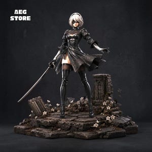 May include: A 2B character figurine with white hair and a black blindfold, wearing a black dress with puff sleeves and thigh-high boots. The figure holds a long sword and stands on a rocky base with small white flowers. Text reads AEG STORE.
