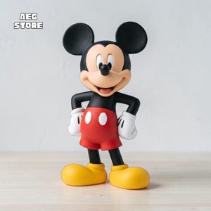 Puede incluir: Una figura de Mickey Mouse con cabeza negra y orejas grandes y redondas, cara color melocotón, torso negro, guantes blancos, pantalones cortos rojos con dos botones ovalados blancos y zapatos amarillos grandes. El texto AEG STORE es visible.