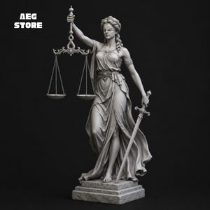 Puede incluir: Una estatua gris de la Dama de la Justicia sobre una base rectangular. La figura lleva una venda en los ojos y una túnica drapeada, sostiene una balanza elevada en la mano derecha y una espada apuntando hacia abajo en la izquierda. El texto AEG STORE es visible.