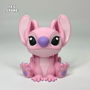 May include: A pink figurine of the character Angel from the Lilo & Stitch franchise. The figurine has large ears, a purple nose, and black eyes. The figurine is sitting down with its hands and feet visible. The text "AEG STORE" is in the top left corner.