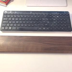 May include: A black HP wireless keyboard with white trim sits above a dark brown wooden wrist rest. The keyboard has black keys and a standard layout. The wrist rest is rectangular with rounded edges. A silver mouse is to the right.