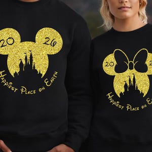K&ouml;nnte beinhalten: Schwarze Sweatshirts mit goldglitzernden Mickey Mouse- und Minnie Mouse-Designs. Das Mickey Mouse-Design hat die Zahlen "20" und "26" und die Worte "Happiest Place on Earth". Das Minnie Mouse-Design hat eine Schleife.