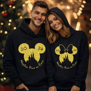 K&ouml;nnte beinhalten: Schwarze Kapuzenpullover mit goldenen Glitzer-Mickey-Mouse- und Minnie-Mouse-Designs. Die Sweatshirts zeigen die Zahlen "2026" und den Text "Happiest Place on Earth".