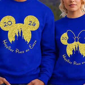 K&ouml;nnte beinhalten: K&ouml;nigsblaue Sweatshirts mit goldenen Glitzer-Mickey-Mouse-Kopf-Designs. Das Design beinhaltet die Zahlen "20" und "26", eine Schlosssilhouette und den Text "Happiest Place on Earth". Ein Sweatshirt hat eine Schleife.