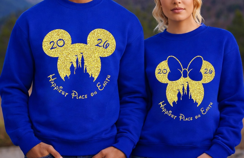 K&ouml;nnte beinhalten: K&ouml;nigsblaue Sweatshirts mit goldenen Glitzer-Mickey-Mouse-Kopf-Designs. Das Design beinhaltet die Zahlen "20" und "26", eine Schlosssilhouette und den Text "Happiest Place on Earth". Ein Sweatshirt hat eine Schleife.