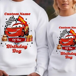 May include: White sweatshirt featuring a red race car graphic with the text "Custom Name Of The Birthday Boy". The design includes checkered flags, a birthday cake, and gift boxes. The sweatshirt is suitable for a birthday celebration.