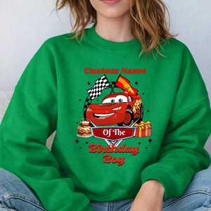 May include: Green sweatshirt featuring a red cartoon race car with the text "Custom Name Of The Birthday Boy." The design includes a birthday cake, wrapped gifts, and checkered flags. The sweatshirt is a solid green color.