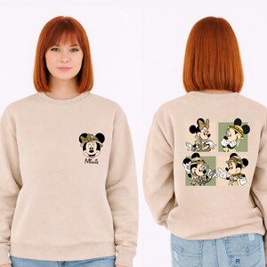 May include: Beige sweatshirt with Minnie Mouse in safari gear on the front and back. The front shows Minnie's face, while the back has four images of Minnie and Mickey Mouse in safari outfits, set against a green backdrop.
