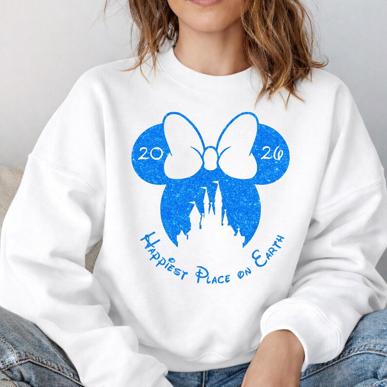 K&ouml;nnte beinhalten: Wei&szlig;es Sweatshirt mit blauem Aufdruck, der eine Schleife, die Zahlen "20" und "20" sowie eine Schloss-Silhouette zeigt. Der Text "Happiest Place on Earth" ist unter dem Schlossbogen angeordnet.