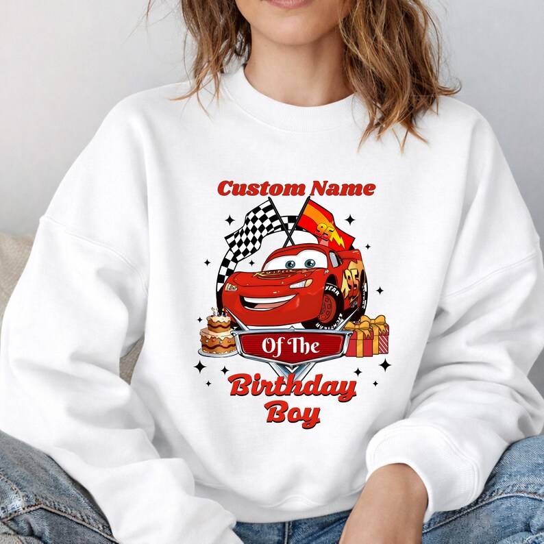 May include: White sweatshirt featuring a red cartoon race car with the text "Custom Name Of The Birthday Boy". The design includes a checkered flag, a birthday cake, and wrapped gifts. The sweatshirt is a comfortable, casual style.