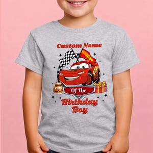 May include: A gray t-shirt featuring a red cartoon race car with the text "Custom Name" above and "Of The Birthday Boy" below. The design includes checkered flags, a birthday cake, and a gift.