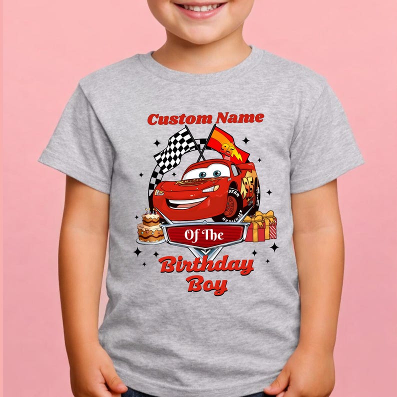 May include: A gray t-shirt featuring a red cartoon race car with the text "Custom Name" above and "Of The Birthday Boy" below. The design includes checkered flags, a birthday cake, and a gift.