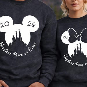 K&ouml;nnte beinhalten: Zwei dunkelgraue Sweatshirts mit wei&szlig;en Grafikdesigns. Eines zeigt einen Mickey Mouse-Kopf mit den Zahlen "20" und "26", eine Schlosssilhouette und den Text "Happiest Place on Earth". Das andere hat einen Minnie Mouse-Kopf mit einer Schleife.