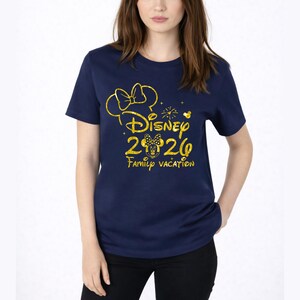 May include: Navy blue t-shirt with a gold glitter design. The design includes a bow, the word "Disney", the numbers "2026", a Minnie Mouse head, and the words "Family Vacation". The shirt is a crew neck style.