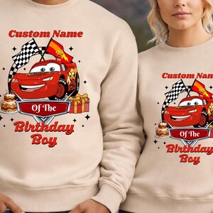 May include: Beige sweatshirt featuring a red cartoon race car with the text "Custom Name" above and "Of The Birthday Boy" below. The design includes checkered flags, a cake, and wrapped gifts.