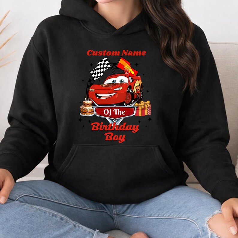 May include: Black hooded sweatshirt featuring a graphic of a red race car with the text "Custom Name Of The Birthday Boy". The design includes checkered flags, a birthday cake, and wrapped gifts.
