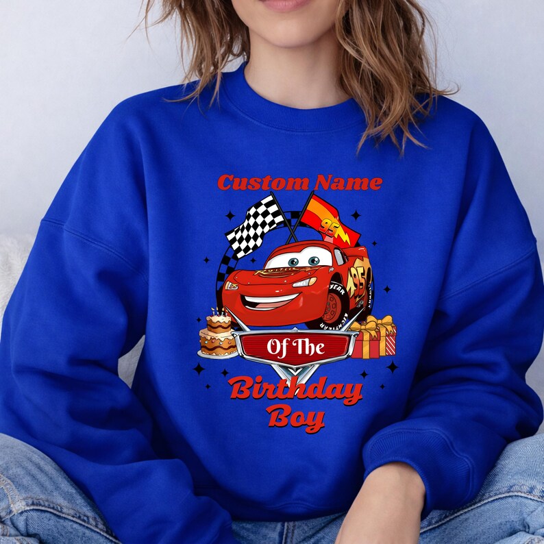 May include: Royal blue sweatshirt featuring a red cartoon race car with the text "Custom Name" above and "Of The Birthday Boy" below. The design includes a checkered flag, a birthday cake, and wrapped gifts.