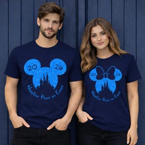 K&ouml;nnte beinhalten: Marineblaue T-Shirts mit einem blauen Glitzerdesign von Mickey Mouse-Ohren und einer Schleife, mit dem Text "2026 Happiest Place on Earth". Ein Mann und eine Frau tragen diese T-Shirts.