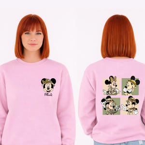 May include: A pale pink sweatshirt with Minnie Mouse graphics. The front displays Minnie in a safari hat, with "Minnie" below. The back features four panels with Minnie in safari attire. The design is a fun, cartoon style.