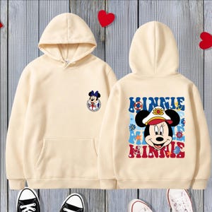May include: Cream-colored hoodie with a front pocket and a hood. The front features a small Minnie Mouse graphic, while the back displays a larger Minnie Mouse design with the name "MINNIE" in blue and red lettering.