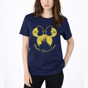 K&ouml;nnte beinhalten: Marineblaues T-Shirt mit einem goldglitzernden Design aus Schleife und Schloss-Silhouette. Der Text "Happiest Place on Earth" und die Zahlen "2020" sind ebenfalls in Gold gehalten. Das Shirt hat einen Rundhalsausschnitt und kurze &Auml;rmel.
