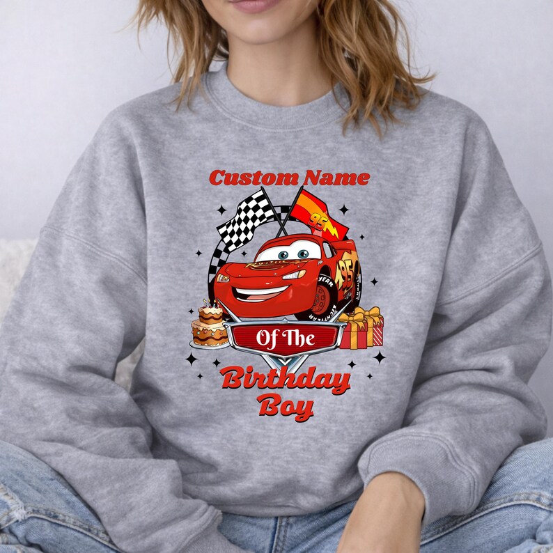 May include: A light gray sweatshirt featuring a red cartoon race car with the text "Custom Name" above and "Of The Birthday Boy" below. The design includes a checkered flag, a birthday cake, and a wrapped gift.