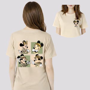 May include: A beige t-shirt with a cartoon design of Mickey and Minnie Mouse in safari outfits. The design is on the back of the shirt, with a smaller version of Minnie Mouse on the front. The shirt has short sleeves.