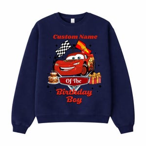May include: Navy blue sweatshirt featuring a red cartoon race car with the text "Custom Name" above and "Of The Birthday Boy" below. The design includes a checkered flag, a birthday cake, and wrapped gifts.