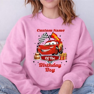 May include: Pink sweatshirt featuring a red cartoon race car with the text "Custom Name" above and "Of The Birthday Boy" below. The design includes a checkered flag, a birthday cake, and a wrapped gift.
