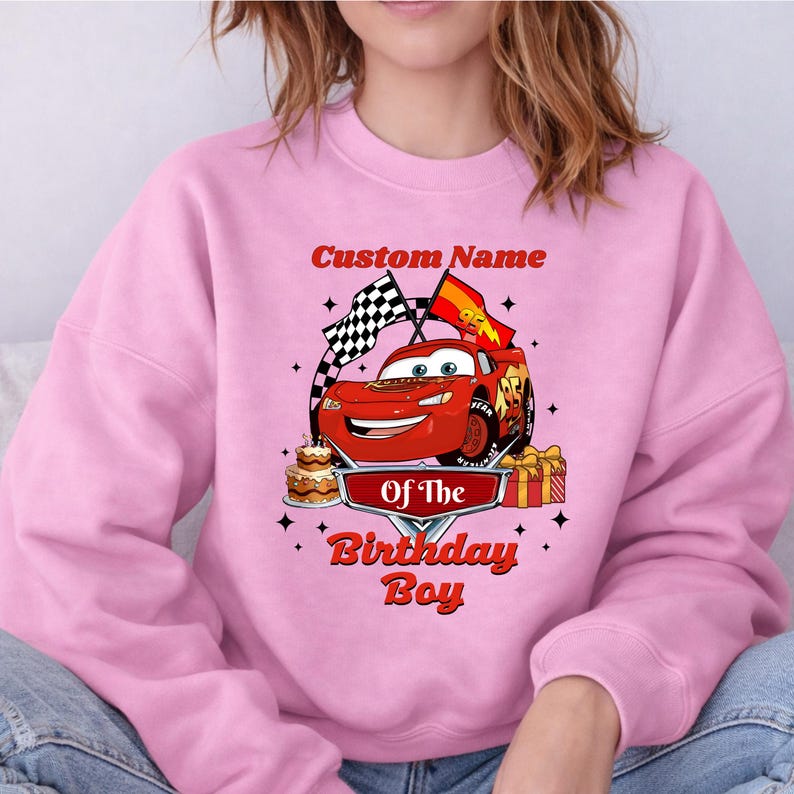 May include: Pink sweatshirt featuring a red cartoon race car with the text "Custom Name" above and "Of The Birthday Boy" below. The design includes a checkered flag, a birthday cake, and a wrapped gift.