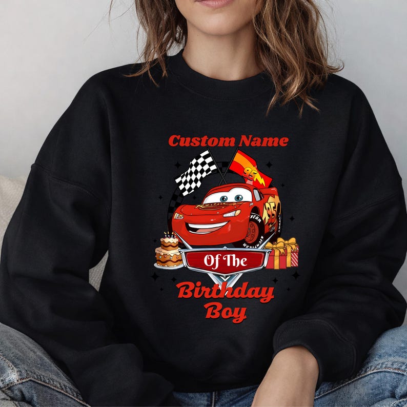 May include: Black sweatshirt featuring a red cartoon race car with the text "Custom Name" above and "Of The Birthday Boy" below. The car has a checkered flag and a birthday cake. A gift box is also visible.