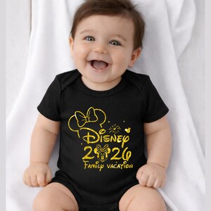 May include: A black baby bodysuit with a gold Minnie Mouse design. The design includes a bow, the word "Disney", the year "2020", and the words "Family Vacation". The baby is smiling and sitting on a white surface.