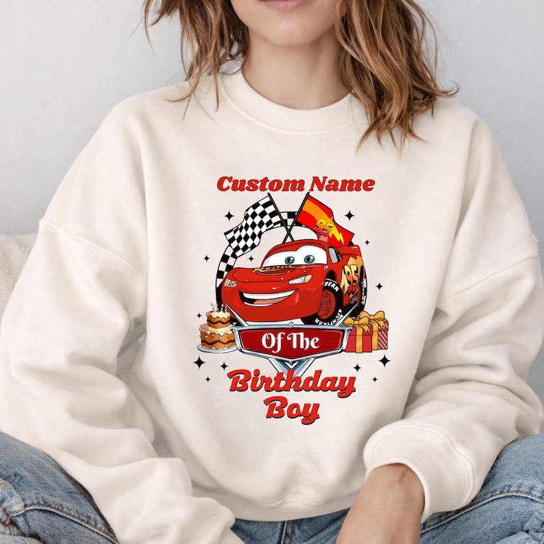 May include: Cream-colored sweatshirt featuring a red cartoon race car with the text "Custom Name Of The Birthday Boy." The design includes checkered flags, a birthday cake, and wrapped gifts. The sweatshirt is a comfortable, casual style.