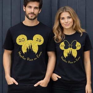 K&ouml;nnte beinhalten: Schwarze T-Shirts mit goldenem Glitzerdesign. Ein Shirt zeigt einen Mickey Mouse-Kopf mit den Zahlen "20" und "26" und dem Text "Happiest Place on Earth". Das andere Shirt hat einen Minnie Mouse-Kopf mit einer Schleife.