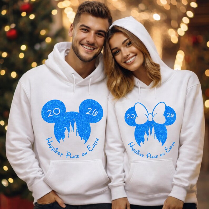 K&ouml;nnte beinhalten: Wei&szlig;e Kapuzenpullover mit blauen Mickey Mouse- und Minnie Mouse-Designs. Jeder Pullover zeigt eine blaue Silhouette eines Schlosses, die Zahlen "20" und "26" und den Text "Happiest Place on Earth".