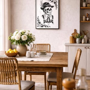 Puede incluir: Una impresi&oacute;n en blanco y negro enmarcada de una calavera con sombrero y tirantes, rodeada de detalles florales. La obra de arte cuelga sobre una mesa de comedor de madera con un jarr&oacute;n de flores blancas, creando un dise&ntilde;o interior r&uacute;stico y art&iacute;stico.