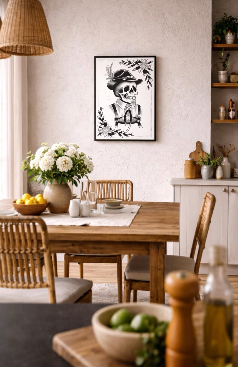 Puede incluir: Una impresi&oacute;n en blanco y negro enmarcada de una calavera con sombrero y tirantes, rodeada de detalles florales. La obra de arte cuelga sobre una mesa de comedor de madera con un jarr&oacute;n de flores blancas, creando un dise&ntilde;o interior r&uacute;stico y art&iacute;stico.
