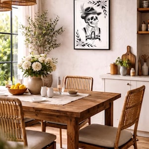Puede incluir: Un comedor con mesa y sillas de madera, una impresi&oacute;n en blanco y negro enmarcada de un esqueleto con vestimenta tradicional y un jarr&oacute;n con flores blancas. La habitaci&oacute;n tiene una paleta de colores neutros.