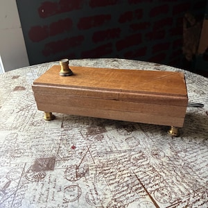 Könnte beinhalten: Eine rechteckige Holzkiste mit einem spulenförmigen Messingknopf und Messingfüßen. Die Kiste ist aus hellbraunem Holz gefertigt und hat einen kleinen Metallgriff an der Seite. Die Kiste steht auf einem Tisch mit Vintage-Buchstabenmuster.
