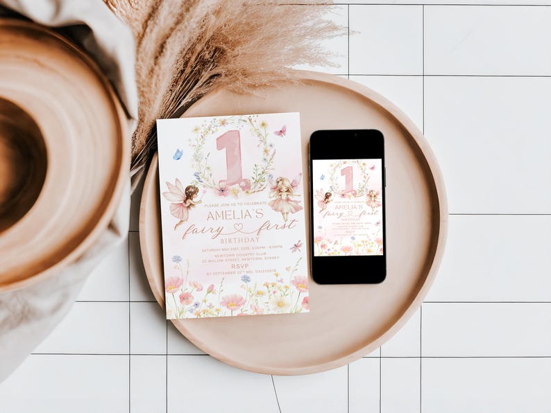 May include: A pink and floral fairy-themed invitation and a smartphone displaying the same design, both on a round tray. The invitation reads "Amelia's fairy first birthday" with fairy illustrations and the number 1.