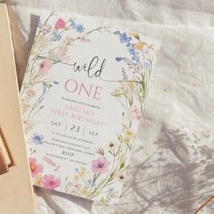 May include: A floral-themed invitation for a first birthday celebration. The card features the text "wild ONE" in a script font, with details about the event, including the date and location, surrounded by a watercolor floral border with pink, blue, and yellow flowers.